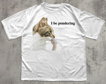May include: White t-shirt featuring a cat graphic in a thinking pose, with the text "I be pondering" in black. The cat has brown and white fur. The shirt is a casual, unisex style.