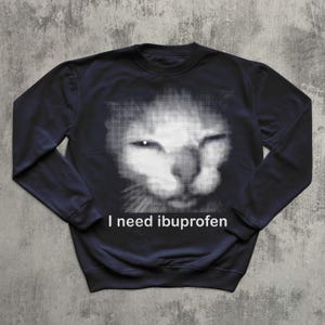 May include: Navy blue sweatshirt featuring a halftone image of a cat's face. The cat's face is white, with the rest of the sweatshirt being navy blue. The text "I need ibuprofen" is printed in white below the cat's face.