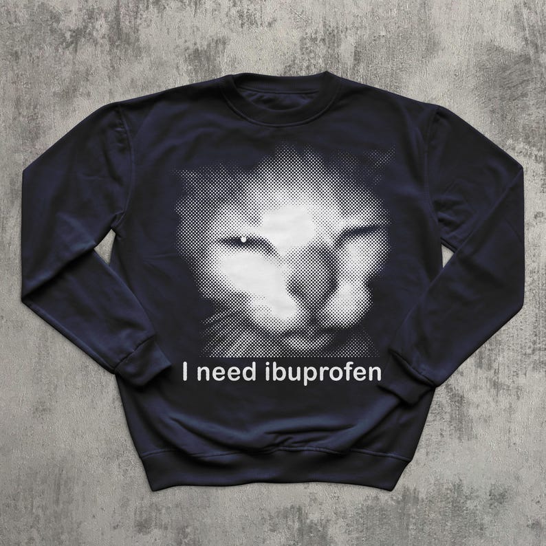 May include: Navy blue sweatshirt featuring a halftone image of a cat's face. The cat's face is white, with the rest of the sweatshirt being navy blue. The text "I need ibuprofen" is printed in white below the cat's face.