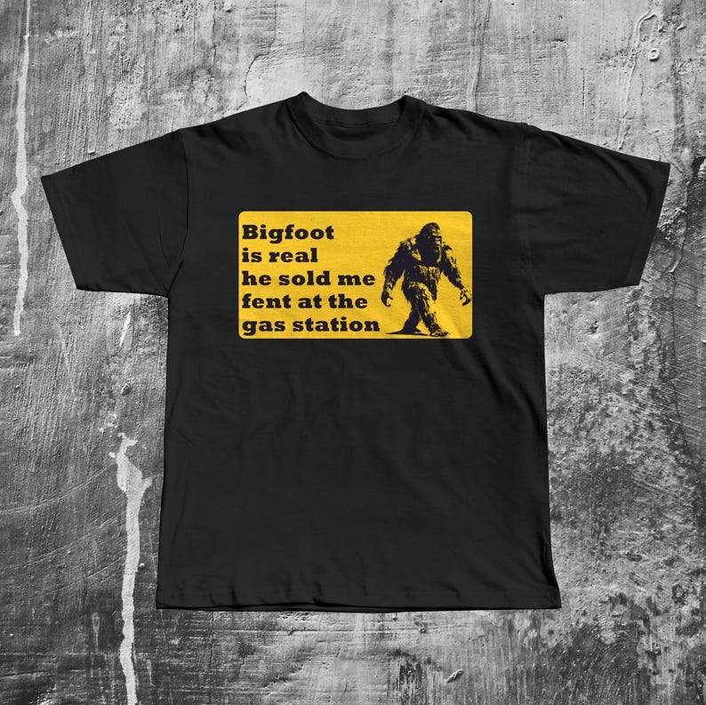 K&ouml;nnte beinhalten: Schwarzes T-Shirt mit einem gelben Schild, auf dem steht "Bigfoot is real he sold me fent at the gas station". Auf dem Schild ist eine Silhouette von Bigfoot abgebildet.
