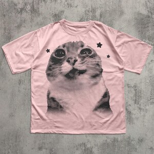 May include: A light pink t-shirt featuring a large, halftone image of a cat's face. The cat's face is in black and white, with three black star shapes around it. The t-shirt has short sleeves and a crew neck.