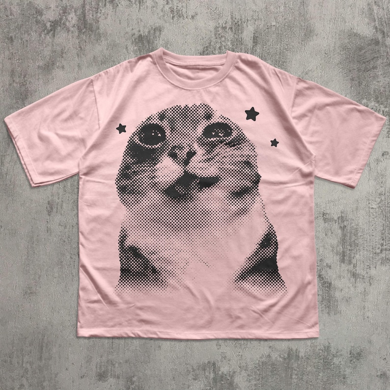 May include: A light pink t-shirt featuring a large, halftone image of a cat's face. The cat's face is in black and white, with three black star shapes around it. The t-shirt has short sleeves and a crew neck.