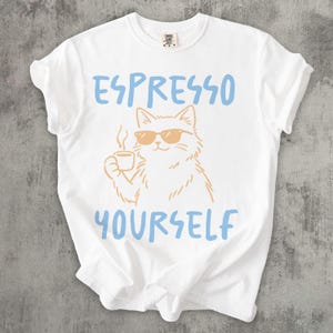 May include: White t-shirt with the text "ESPRESSO YOURSELF" in light blue. The shirt features a cartoon cat wearing sunglasses and holding a coffee cup. The cat and coffee cup are in a light brown color.