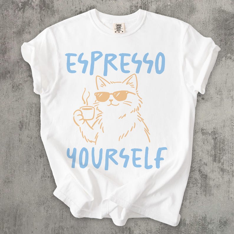 May include: White t-shirt with the text "ESPRESSO YOURSELF" in light blue. The shirt features a cartoon cat wearing sunglasses and holding a coffee cup. The cat and coffee cup are in a light brown color.