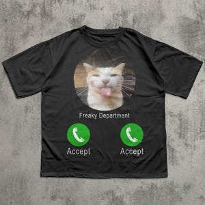 以下が含まれることがあります： 黒いTシャツ。舌を出している猫が円の中に描かれています。「Freaky Department」の文字が、2つの緑色の「Accept」ボタンの上にあります。ユーモラスなデザインです。