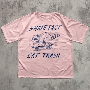 May include: A light pink t-shirt featuring a raccoon skateboarding. The raccoon is illustrated in navy blue, with the words "SKATE FAST" above and "EAT TRASH" below the raccoon. The shirt has short sleeves and a crew neck.