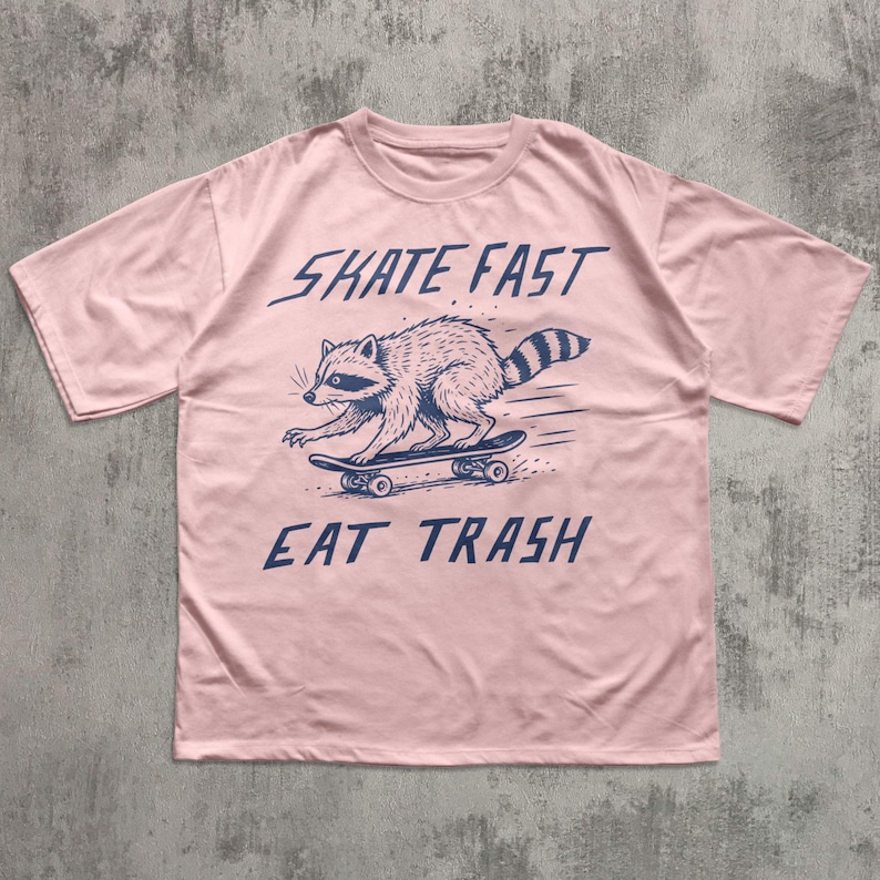 May include: A light pink t-shirt featuring a raccoon skateboarding. The raccoon is illustrated in navy blue, with the words "SKATE FAST" above and "EAT TRASH" below the raccoon. The shirt has short sleeves and a crew neck.