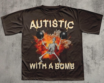Puede incluir: Camiseta marrón con la frase "AUTISTIC WITH A BOMB". El diseño muestra un esqueleto sosteniendo dos bombas negras, con un fondo de llamas y explosión.