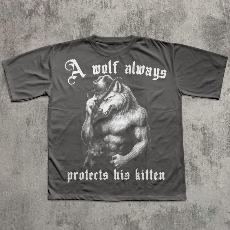 May include: Dark gray t-shirt featuring a muscular wolf wearing a cowboy hat. The wolf illustration is in black and white. The text "A wolf always protects his kitten" is in white, gothic-style font.