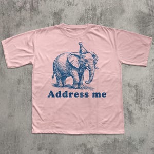 May include: A light pink t-shirt featuring a blue illustration of an elephant wearing a party hat. The text "Address me" is printed below the elephant in blue. The shirt has short sleeves and a crew neck.