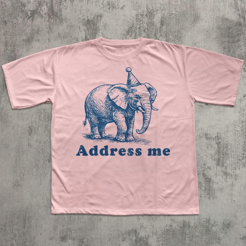 May include: A light pink t-shirt featuring a blue illustration of an elephant wearing a party hat. The text "Address me" is printed below the elephant in blue. The shirt has short sleeves and a crew neck.