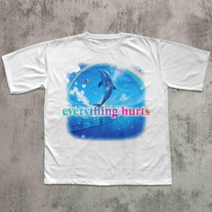 May include: White t-shirt featuring a dolphin leaping from blue waves under a rainbow. The design includes clouds, hearts, and the text "everything hurts" in a colorful gradient. A casual, graphic tee.