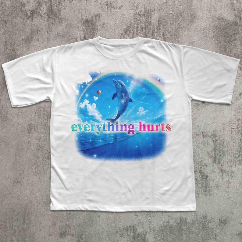 May include: White t-shirt featuring a dolphin leaping from blue waves under a rainbow. The design includes clouds, hearts, and the text "everything hurts" in a colorful gradient. A casual, graphic tee.