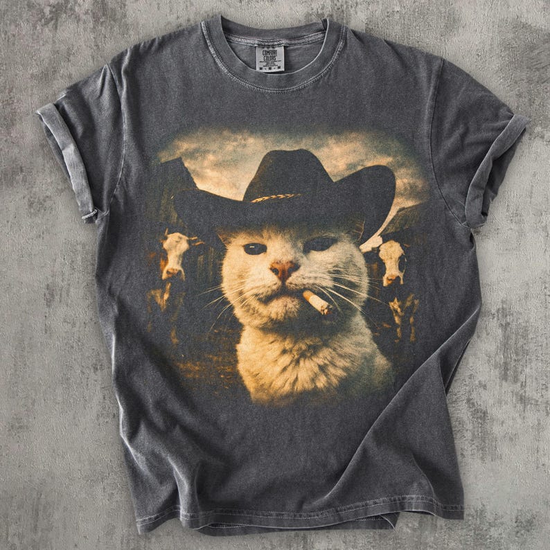 May include: Dark gray t-shirt featuring a white cat wearing a cowboy hat and smoking a cigarette. The cat is in front of a background of cows and a cloudy sky. The shirt has rolled-up sleeves.