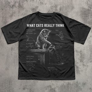 May include: A black t-shirt featuring a white graphic of a cat pushing a glass off a table, surrounded by physics equations and the text "WHAT CATS REALLY THINK". The design includes diagrams for projectile motion, force, and kinetic energy.