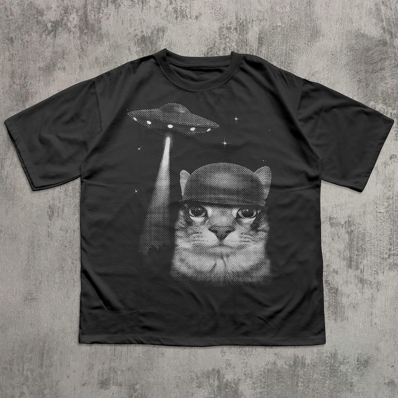 K&ouml;nnte beinhalten: Schwarzes T-Shirt mit einem Halbton-Grafikmotiv einer Katze mit Helm, die von einem UFO angestrahlt wird. Das Design umfasst Sterne und einen Lichtstrahl. Das T-Shirt hat einen Rundhalsausschnitt und kurze &Auml;rmel.