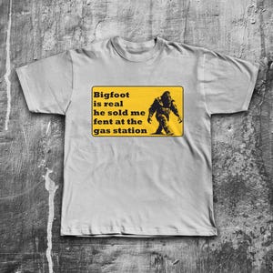 K&ouml;nnte beinhalten: Ein graues T-Shirt mit einem gelben Schild, auf dem steht "Bigfoot is real he sold me fent at the gas station". Auf dem Schild ist eine schwarze Silhouette von Bigfoot abgebildet.