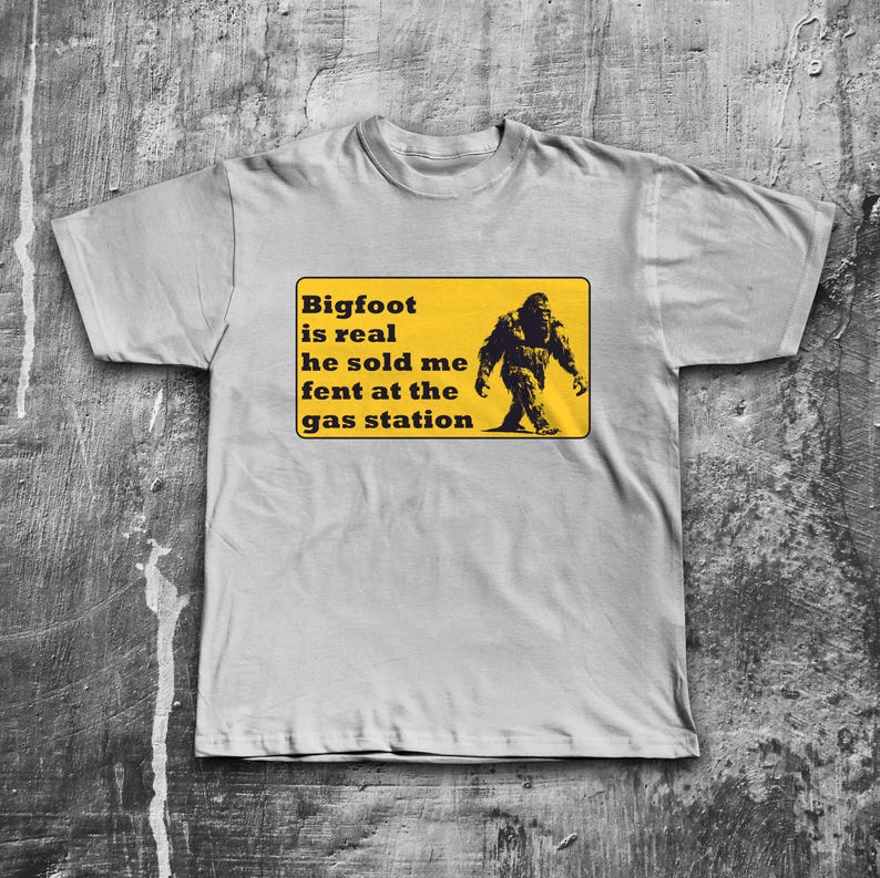 K&ouml;nnte beinhalten: Ein graues T-Shirt mit einem gelben Schild, auf dem steht "Bigfoot is real he sold me fent at the gas station". Auf dem Schild ist eine schwarze Silhouette von Bigfoot abgebildet.