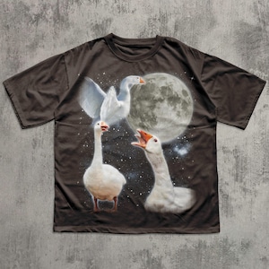 Op de afbeelding: Een donkerbruin t-shirt met korte mouwen en een grafische print van drie witte ganzen in een nachtelijke hemel. Het ontwerp bevat een grote volle maan, verspreide witte sterren en de ganzen in verschillende houdingen tegen de achtergrond.