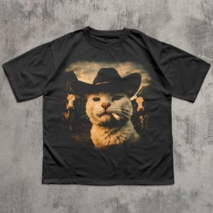 May include: Black t-shirt featuring a humorous graphic of a white cat wearing a cowboy hat and smoking a cigarette. The cat is set against a backdrop of cows and a cloudy sky.