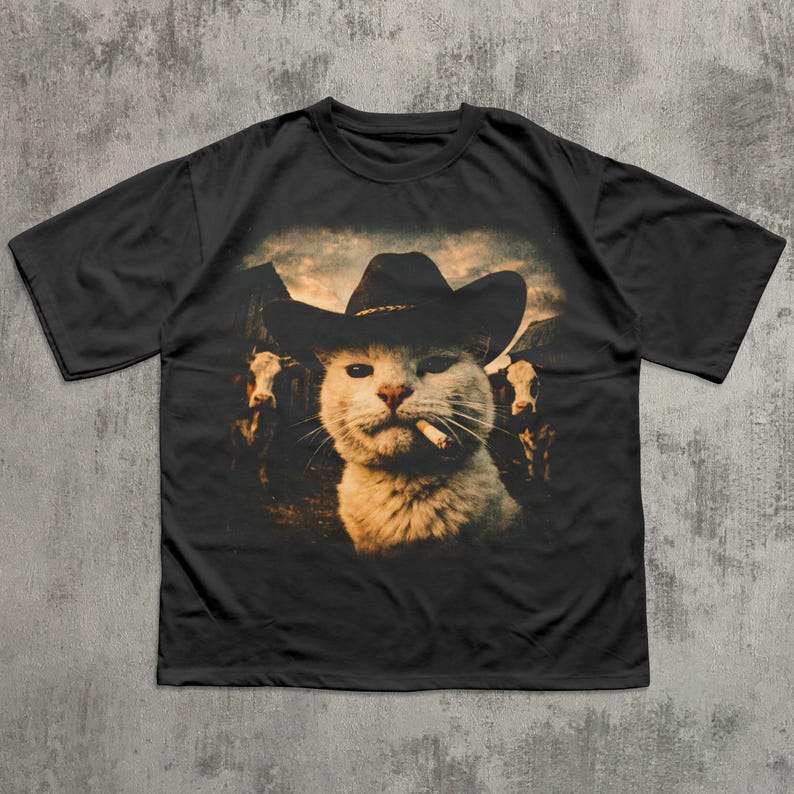 May include: Black t-shirt featuring a humorous graphic of a white cat wearing a cowboy hat and smoking a cigarette. The cat is set against a backdrop of cows and a cloudy sky.