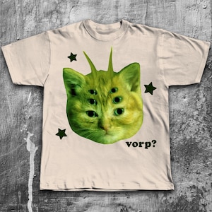 May include: Beige t-shirt featuring a green cat-like creature with multiple eyes and antennae. The design includes green stars and the word "vorp?" in a dark green font. The shirt has a crew neck and short sleeves.