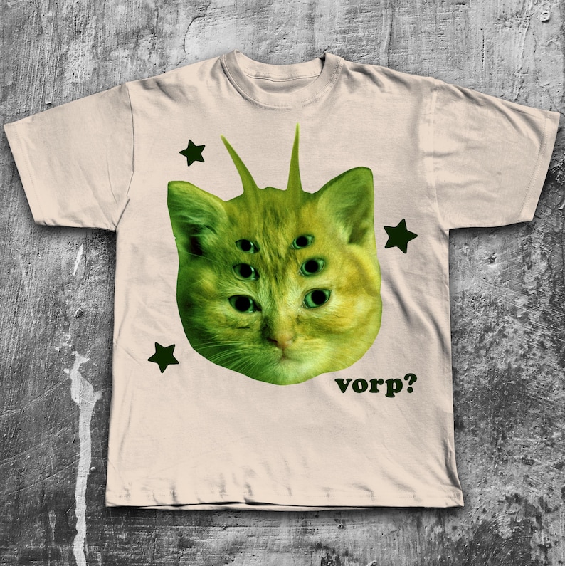 May include: Beige t-shirt featuring a green cat-like creature with multiple eyes and antennae. The design includes green stars and the word "vorp?" in a dark green font. The shirt has a crew neck and short sleeves.