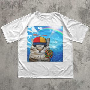 May include: White t-shirt featuring a whimsical graphic of a cat wearing a colorful hat with a propeller and holding a lollipop. The cat is set against a backdrop of a rainbow and a blue sky with clouds. A fun and playful design.