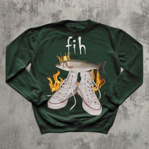 May include: Dark green sweatshirt with a graphic design featuring white high-top trainers, a fish wearing a gold crown, and flames. The word "fih" is written above the fish in white. The sweatshirt is a casual, unisex garment.