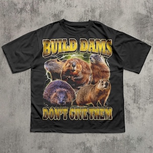 May include: Black t-shirt featuring a graphic of several brown beavers with the text "BUILD DAMS DON'T GIVE THEM" in yellow, outlined in black. The design includes lightning bolts. The shirt has short sleeves and a crew neck.