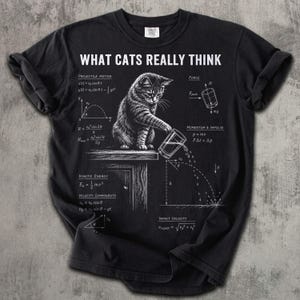 May include: A black t-shirt featuring a white graphic of a cat pushing a glass off a table. The design includes physics equations and diagrams labeled Projectile Motion, Force, Momentum, Kinetic Energy, and Impact Velocity with the text WHAT CATS REALLY THINK.