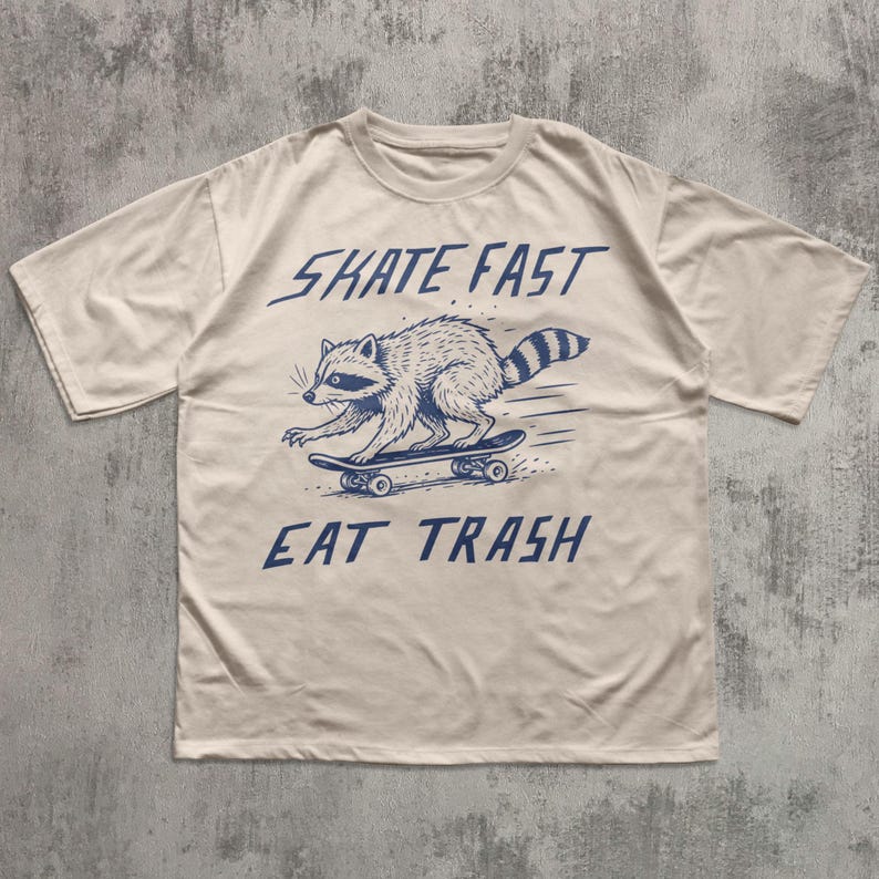 May include: Cream-colored t-shirt featuring a raccoon skateboarding. The raccoon is illustrated in blue, with the words "SKATE FAST" above and "EAT TRASH" below. The design is centered on the front of the shirt.