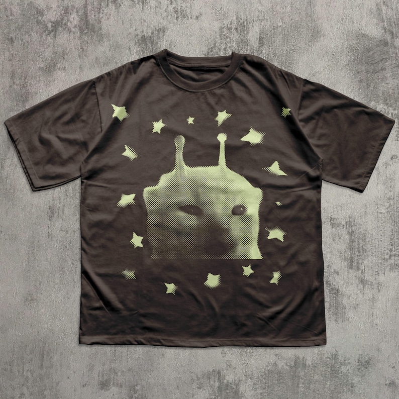 Puede incluir: Camiseta marr&oacute;n con un dise&ntilde;o de cara alien&iacute;gena y estrellas de color verde claro. El gr&aacute;fico es una impresi&oacute;n de semitonos. La camiseta tiene mangas cortas y cuello redondo. La camiseta es de material suave.