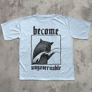 Op de afbeelding: Lichtblauw T-shirt met een zwarte afbeelding van een kat die in een hand bijt, in een zwart rechthoek. De woorden "become" en "ungovernable" staan in gotische letters boven en onder de afbeelding.