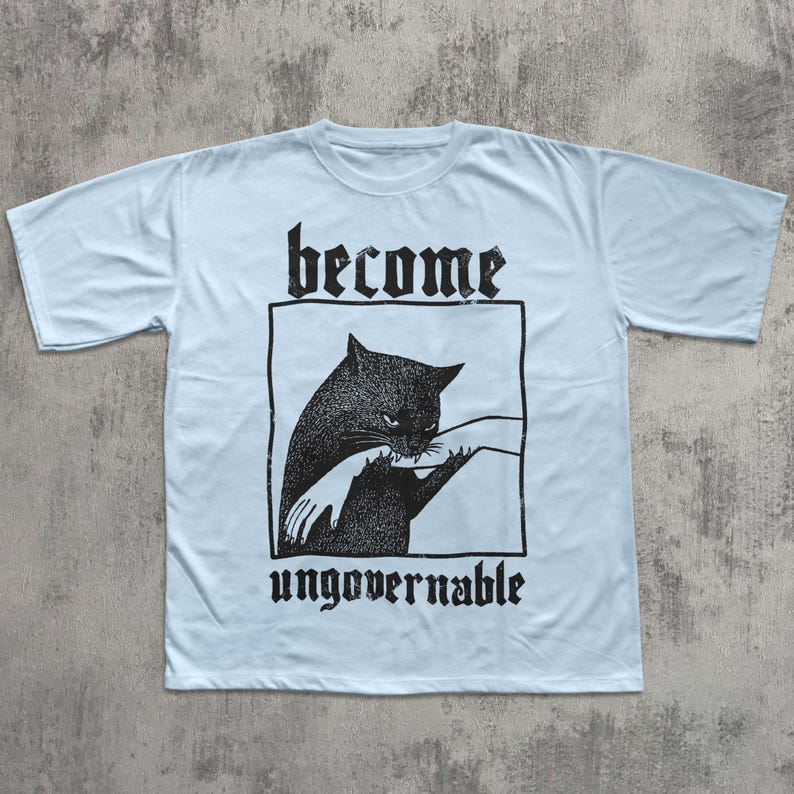 Op de afbeelding: Lichtblauw T-shirt met een zwarte afbeelding van een kat die in een hand bijt, in een zwart rechthoek. De woorden "become" en "ungovernable" staan in gotische letters boven en onder de afbeelding.