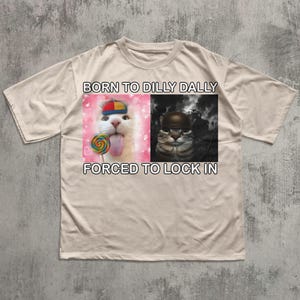 May include: Beige t-shirt featuring a split graphic. The left side shows a white cat with a lollipop and the text "BORN TO DILLY DALLY". The right side shows a cat in a helmet with the text "FORCED TO LOCK IN".