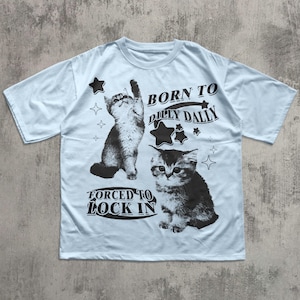 May include: Pale blue t-shirt with two monochrome cat graphics and star designs. The shirt displays the text "BORN TO DILLY DALLY" and "FORCED TO LOCK IN" in black lettering.