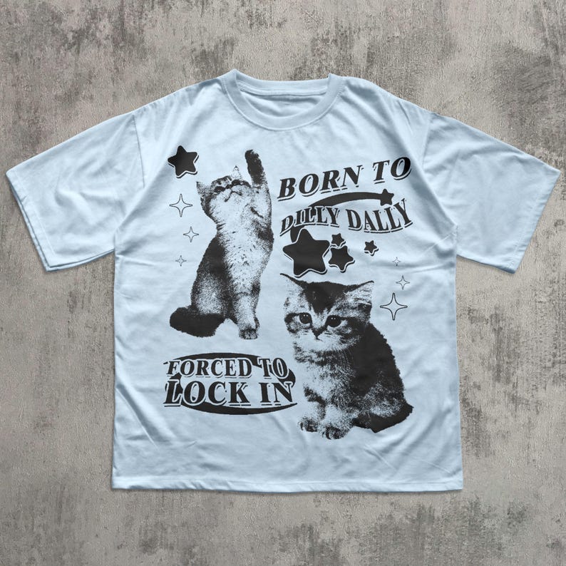 May include: Pale blue t-shirt with two monochrome cat graphics and star designs. The shirt displays the text "BORN TO DILLY DALLY" and "FORCED TO LOCK IN" in black lettering.