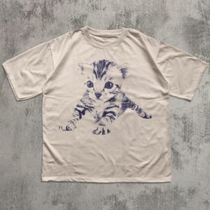 K&ouml;nnte beinhalten: Cremefarbenes T-Shirt mit einer blau-wei&szlig;en Illustration eines K&auml;tzchens. Die Grafik zeigt das K&auml;tzchen in einer spielerischen Pose, mit ausgestreckten Pfoten. Das Shirt hat kurze &Auml;rmel und einen Rundhalsausschnitt.
