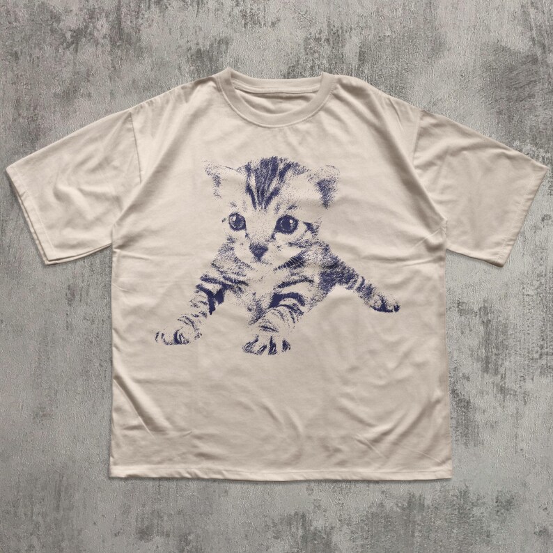 K&ouml;nnte beinhalten: Cremefarbenes T-Shirt mit einer blau-wei&szlig;en Illustration eines K&auml;tzchens. Die Grafik zeigt das K&auml;tzchen in einer spielerischen Pose, mit ausgestreckten Pfoten. Das Shirt hat kurze &Auml;rmel und einen Rundhalsausschnitt.