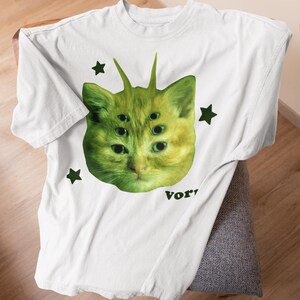 May include: White t-shirt featuring a green cat-like creature with multiple eyes and antennae, surrounded by black star shapes. The word "Vor" is printed in black at the bottom of the design. The shirt is made of a soft, light-colored fabric.