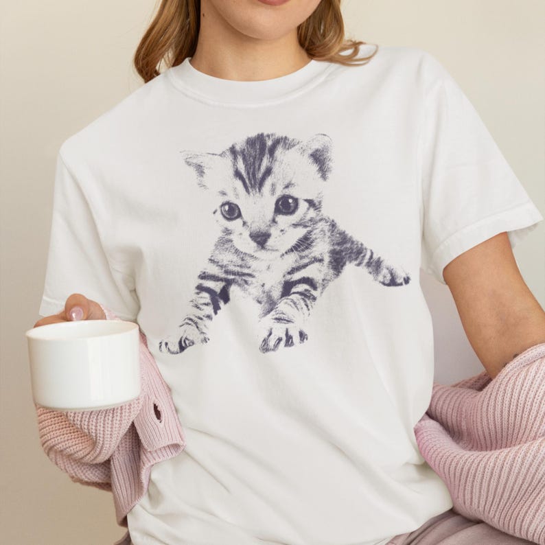 K&ouml;nnte beinhalten: Wei&szlig;es T-Shirt mit einer detaillierten Illustration eines grau-schwarzen K&auml;tzchens. Das K&auml;tzchen wird in einer verspielten Pose dargestellt. Das Shirt besteht aus einem weichen Material. Die Person h&auml;lt einen wei&szlig;en Becher und tr&auml;gt einen rosa Pullover.