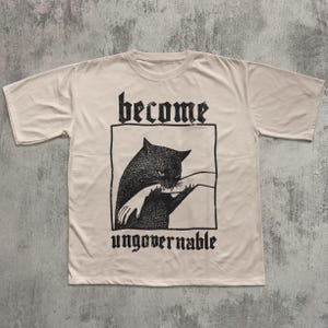 Op de afbeelding: Cr&egrave;mekleurig T-shirt met een zwarte afbeelding van een kat die in een hand bijt, omlijnd in een rechthoek. De woorden "become" en "ungovernable" staan boven en onder de afbeelding in een gotisch lettertype.
