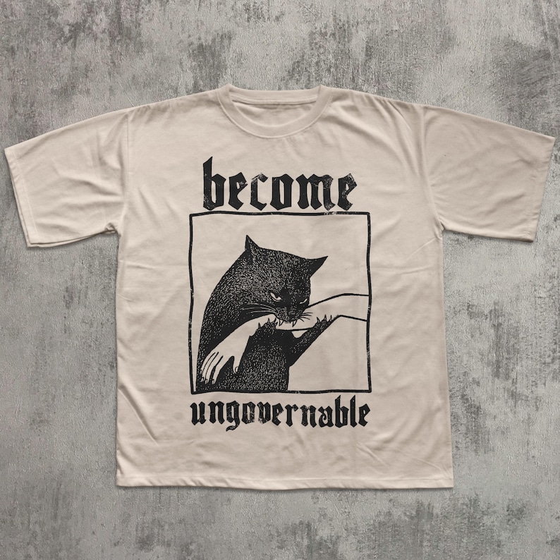 Op de afbeelding: Cr&egrave;mekleurig T-shirt met een zwarte afbeelding van een kat die in een hand bijt, omlijnd in een rechthoek. De woorden "become" en "ungovernable" staan boven en onder de afbeelding in een gotisch lettertype.