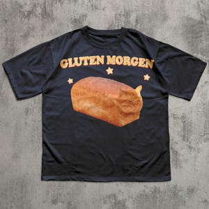 Può includere: T-shirt nero con la scritta "GLUTEN MORGEN" in arancione, sopra una pagnotta a forma di gatto. Il design include tre stelle arancioni. La maglietta è realizzata in un materiale morbido.