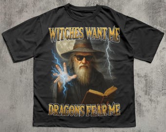 Może przedstawiać: Czarny t-shirt z wizerunkiem czarodzieja w okularach przeciwsłonecznych, kapeluszu i długiej brodzie. Czarodziej trzyma książkę i rzuca zaklęcie z niebieskimi błyskawicami. Tekst "Witches Want Me, Dragons Fear Me" jest nadrukowany.