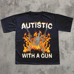 May include: Black t-shirt featuring a skeleton holding two guns, set against a backdrop of flames. The text "AUTISTIC" is arched above the skeleton, with "WITH A GUN" below, all in white lettering. The shirt has a crew neck and short sleeves.