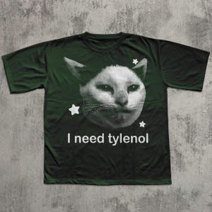 May include: Dark green t-shirt featuring a white, pixelated cat face with star accents. The text "I need tylenol" is printed below the cat image. The shirt is made of a soft material.