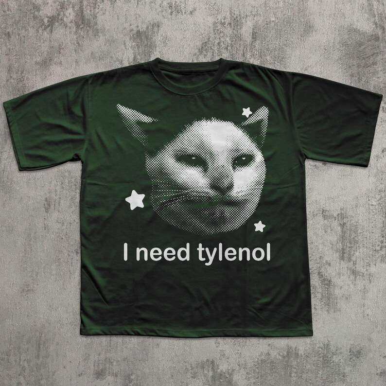 May include: Dark green t-shirt featuring a white, pixelated cat face with star accents. The text "I need tylenol" is printed below the cat image. The shirt is made of a soft material.