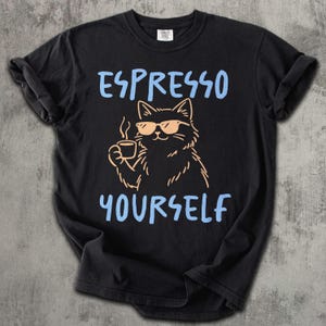 May include: Black t-shirt with the phrase "ESPRESSO YOURSELF" in light blue. A cartoon cat wearing sunglasses and holding a coffee cup is in the center. The cat is drawn in a light orange color.